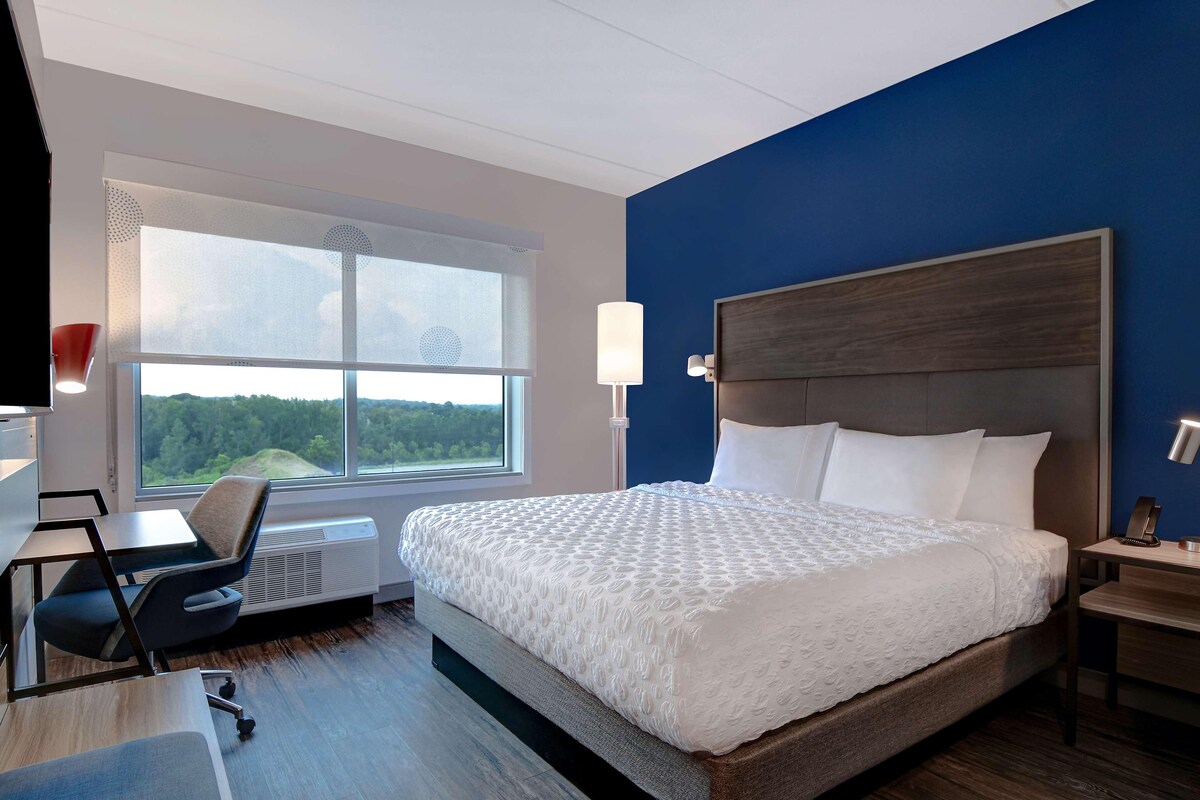 A hotel room with a large bed, a desk, a chair, a window with a view of trees, and a blue accent wall.