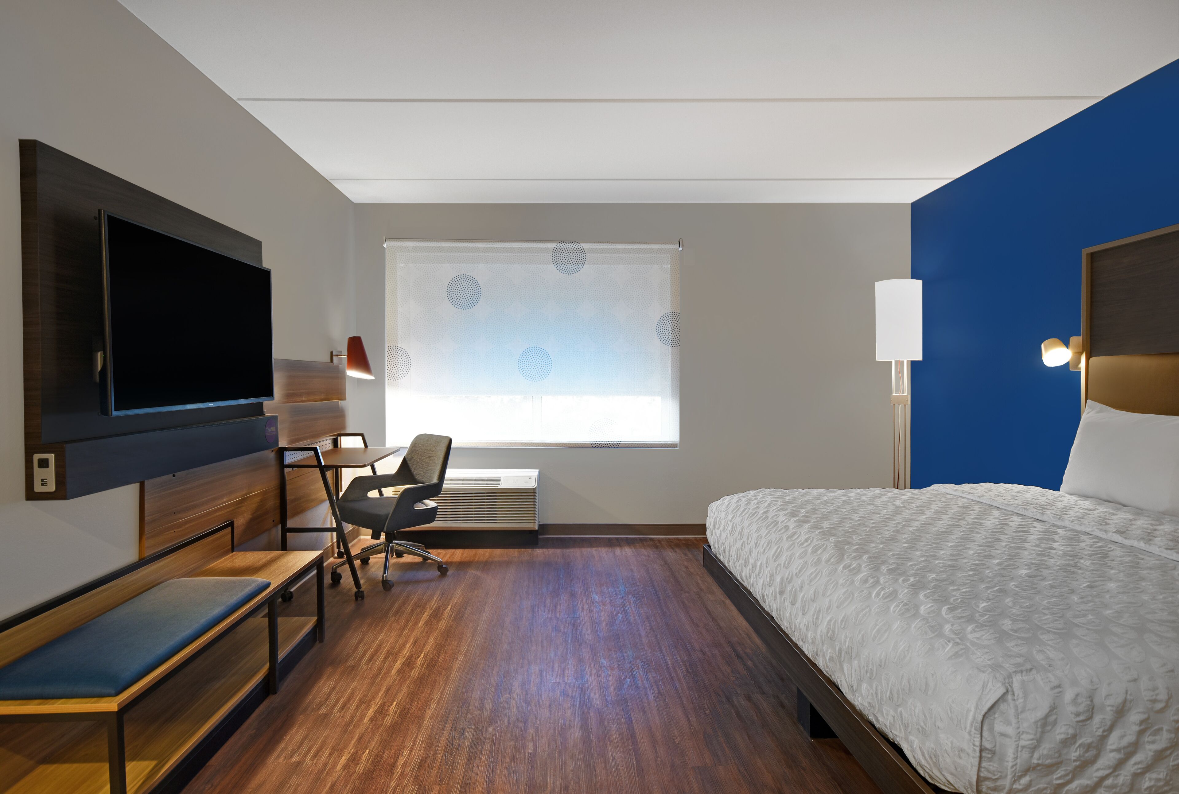 Una habitación de hotel con cama, escritorio, silla, televisión y ventana con persianas.
