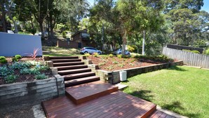 A backyard with a wooden deck, a grassy lawn, and a garden area with plants and mulch. - "Happy Ours" - Walking Distance to Shops & Beach (AVOCA BEACH)