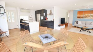 A modern living room with a glass dining table surrounded by wicker chairs, a kitchen area with dark cabinetry, and a large window with a view of the sea. - "Happy Ours" - Walking Distance to Shops & Beach (AVOCA BEACH)