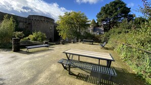 Une cour avec une table de pique-nique et des bancs, entourée de verdure et bordée d’un bâtiment historique en pierre.