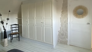 Une pièce avec un plancher en bois peint en blanc, une grande armoire blanche, un mur en pierre et un miroir rond placé au-dessus d’une porte.