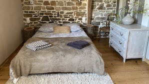 Une chambre avec un mur en pierre, un lit recouvert d’une couverture grise, une commode en bois et un vase avec des plantes.
