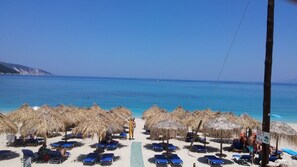 A beach with thatched umbrellas and sun loungers, a clear blue sea, and a distant sailboat.