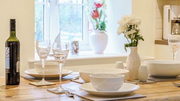 Een gedekte eettafel met witte servies, glazen en een fles wijn, met uitzicht door een raam en bloemen op de achtergrond.