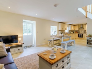 A modern living room with a TV, sofa, and coffee table, a dining area with benches, and a kitchen with wooden cabinets and a stove.