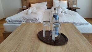 A neatly made bed with white linens and towels, a wooden headboard, and a tray with two bottles and glasses on a wooden table.