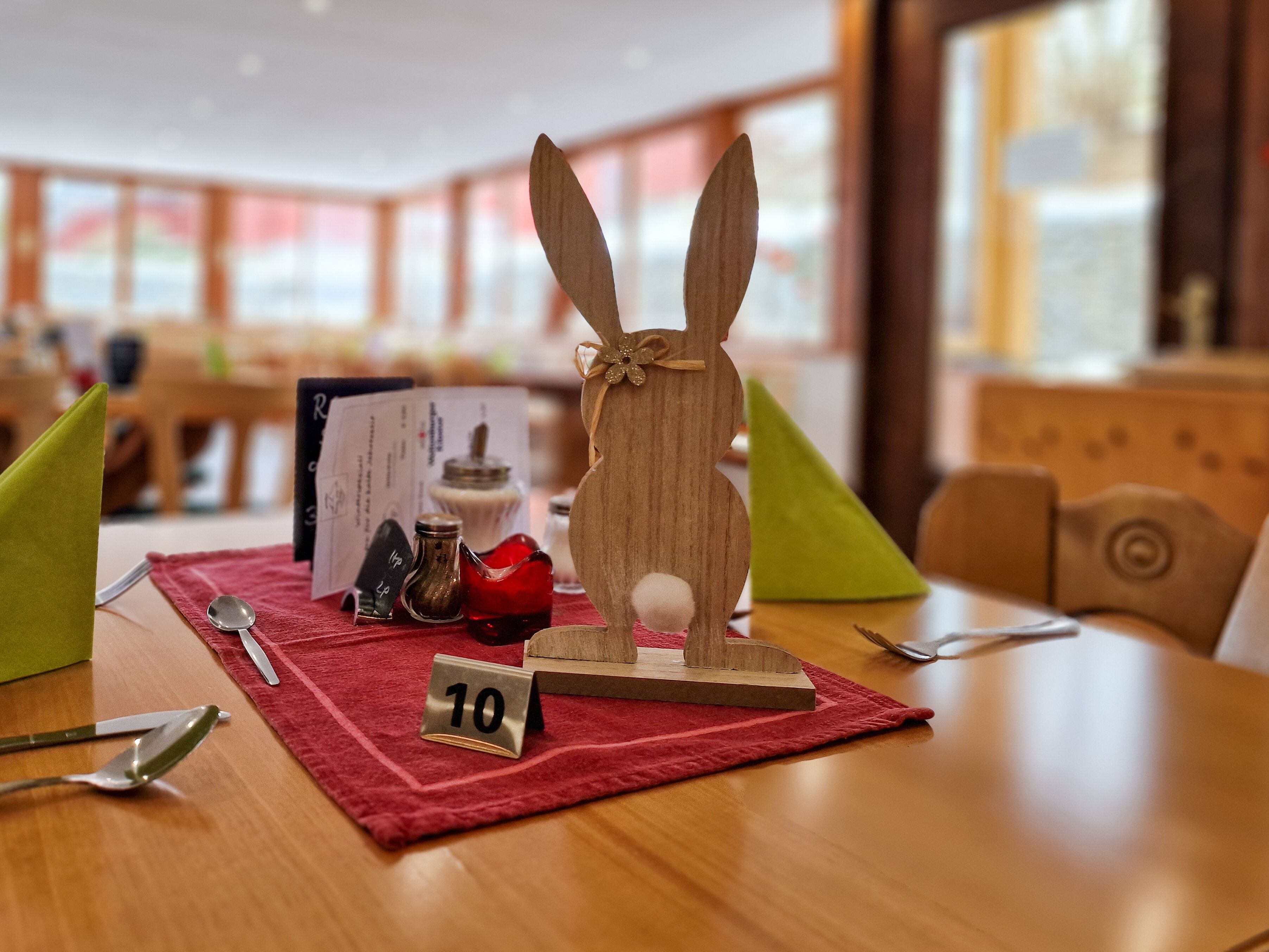 Uma figura de coelho de madeira sobre uma mesa com um jogo americano vermelho, uma colher, um saleiro, um copo de vidro vermelho e uma pequena placa com o número 10.