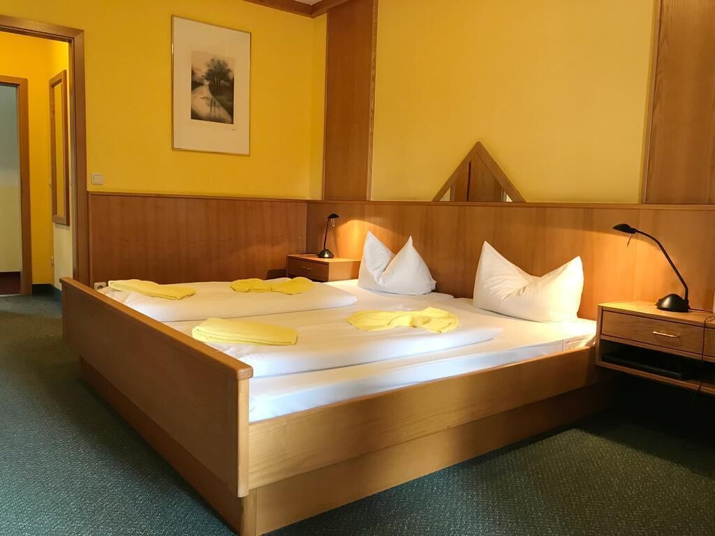 Una cama bien hecha con ropa de cama blanca y amarilla, un cabecero de madera, una mesita de noche con una lámpara y un cuadro en la pared.