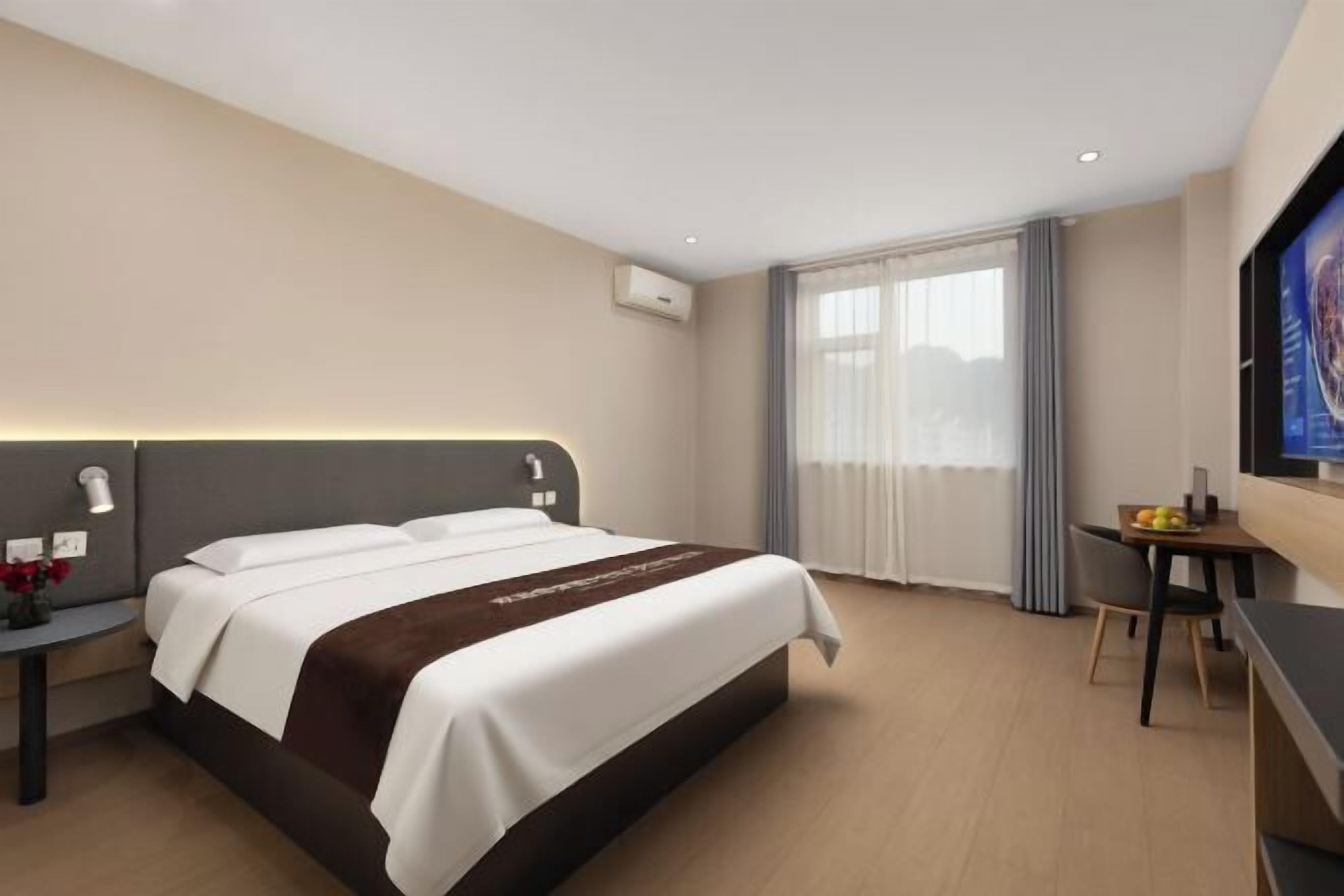 Ett hotellrum med en säng, ett sängbord, en lampa, en luftkonditioneringsanläggning, ett fönster med gardiner och ett litet bord med frukt.