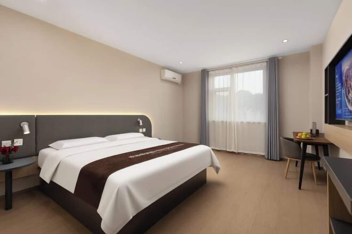 A hotel room with a bed, bedside table, lamp, air conditioning unit, window with curtains, and a small table with fruits.