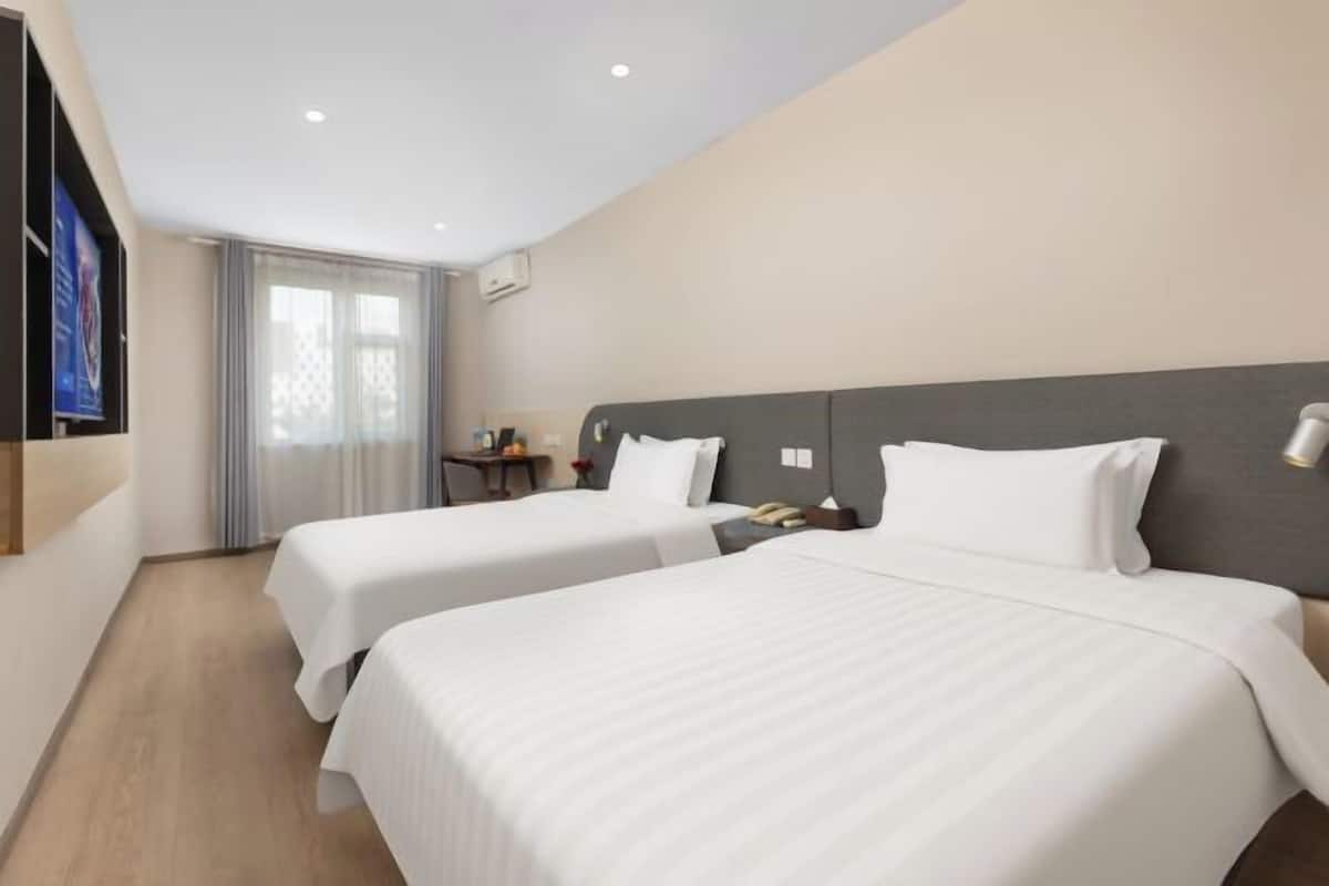 A hotel room with two beds, a TV mounted on the wall, a desk with a chair, and a window with curtains.