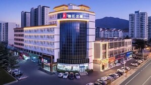 A modern hotel with a prominent curved glass facade, multiple floors, and a parking lot with cars. - Jinjiang Inn (Weihai High-speed Railway North Station Oulefang Commercial Street Hotel) (Weihai)