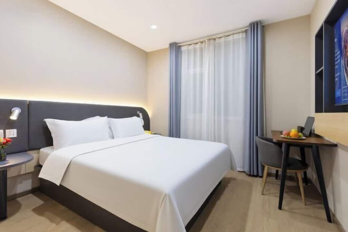 A hotel room with a bed, a desk with a chair, a small table with fruits, and a window with curtains.