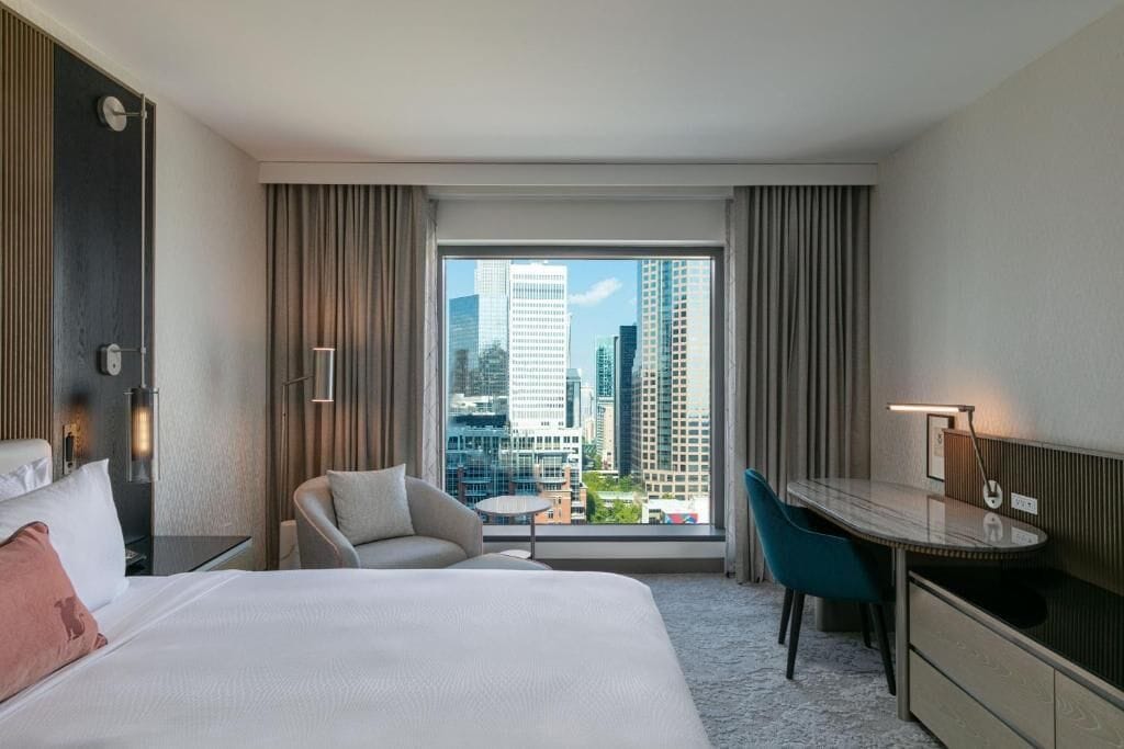 A hotel room with a bed, a desk, a chair, and a view of city buildings.