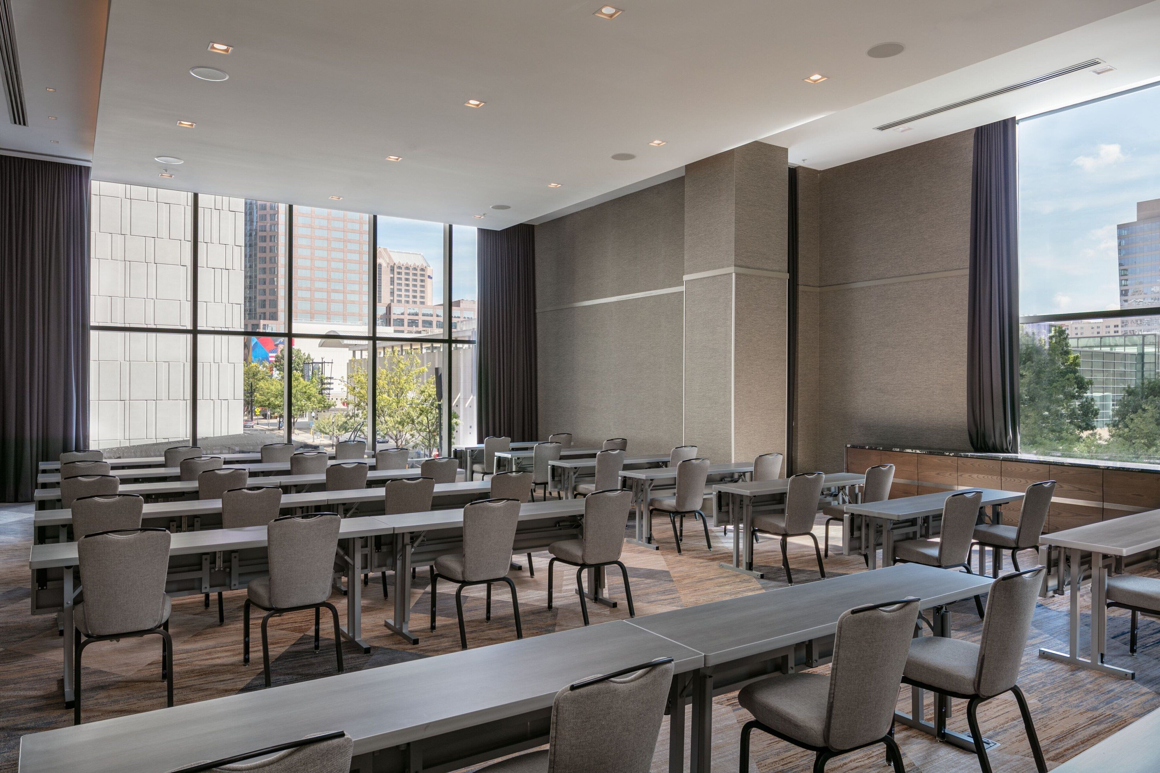 Un salón de conferencias con varias mesas y sillas, ventanas amplias con cortinas y vistas a edificios.