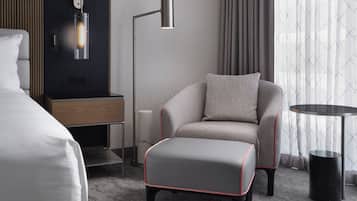 Una habitación de hotel moderna con un sillón tapizado gris, un otoman a juego, una mesita de noche con lámpara y un ventanal con cortinas.