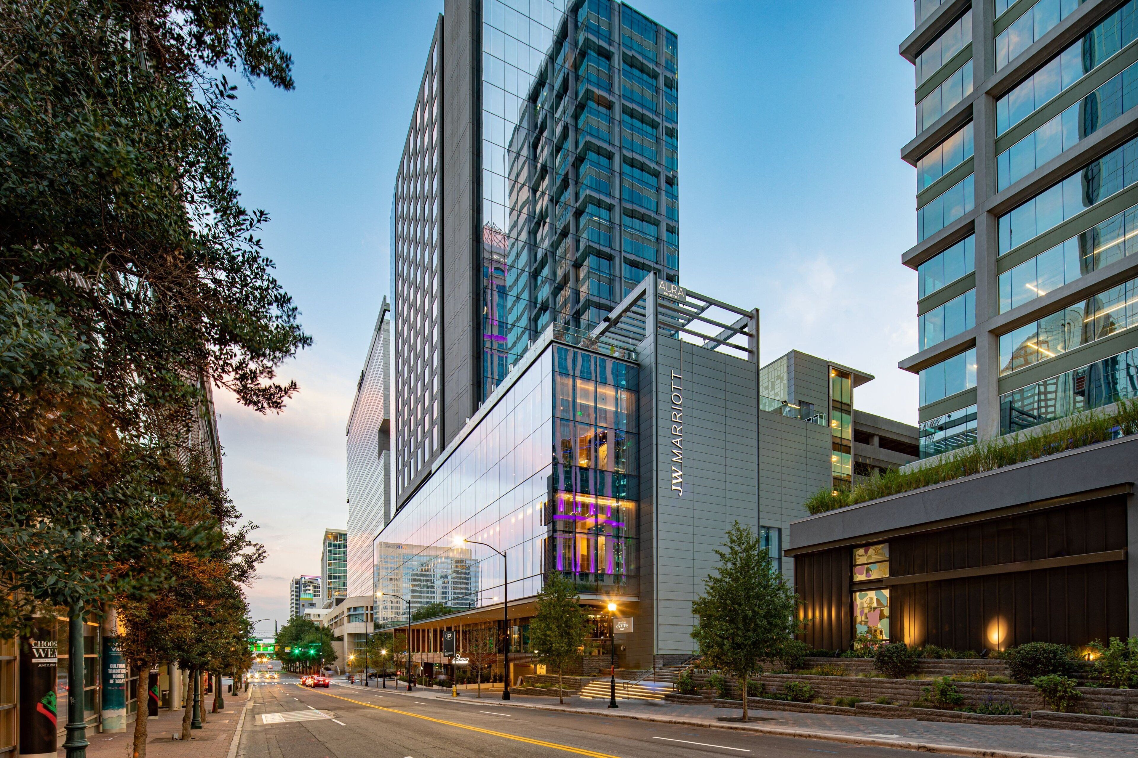 Un hotel moderno con fachada de vidrio, un letrero llamativo de JW Marriott y vista a la calle con árboles y otros edificios.