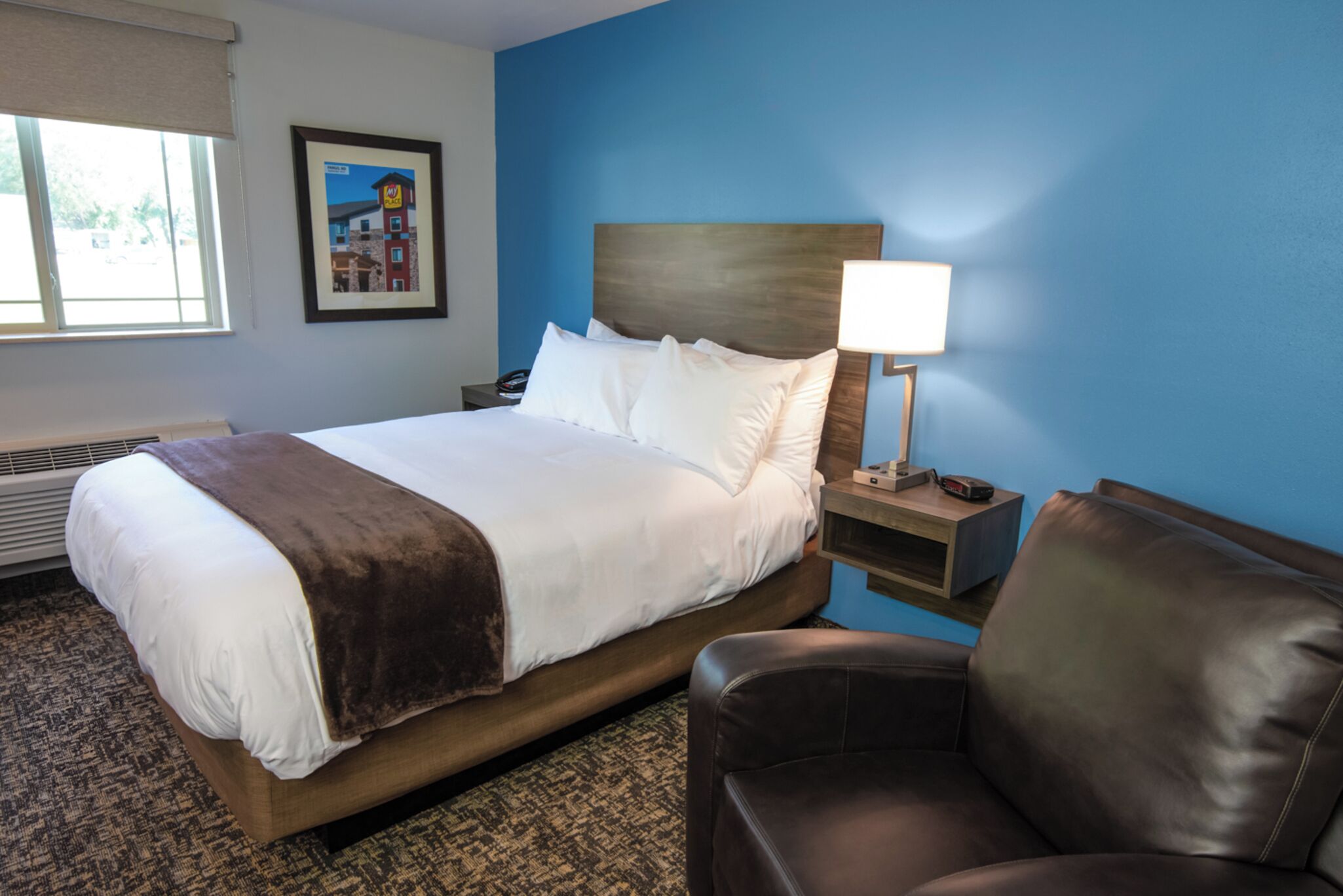 Une chambre d’hôtel avec un grand lit, une table de chevet avec une lampe, un fauteuil en cuir marron et un tableau encadré au mur.