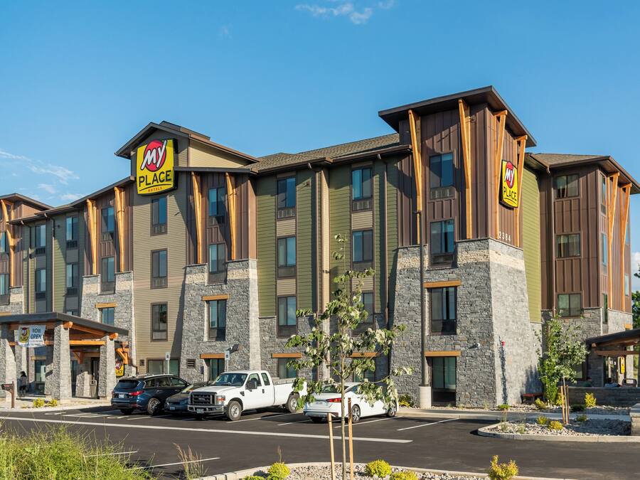 Un hotel de varios pisos con un letrero que dice 'My Place', fachada de piedra y madera, y un estacionamiento con varios automóviles.
