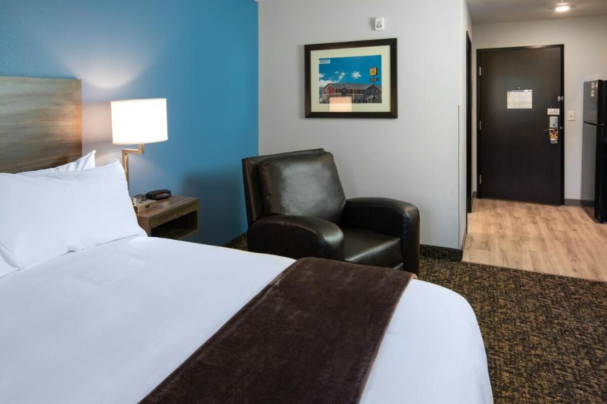 A hotel room with a bed, a chair, a desk with a lamp, a framed picture on the wall, and a door.