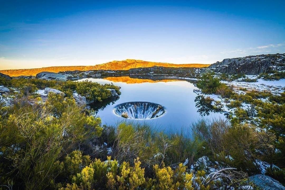 Eine natürliche Landschaft mit einem kreisförmigen Wasserspiel, umgeben von Vegetation und felsigem Gelände.