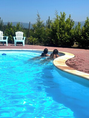 Ein Schwimmbecken mit zwei Personen im Wasser, umgeben von viel Grün und blauem Himmel.
