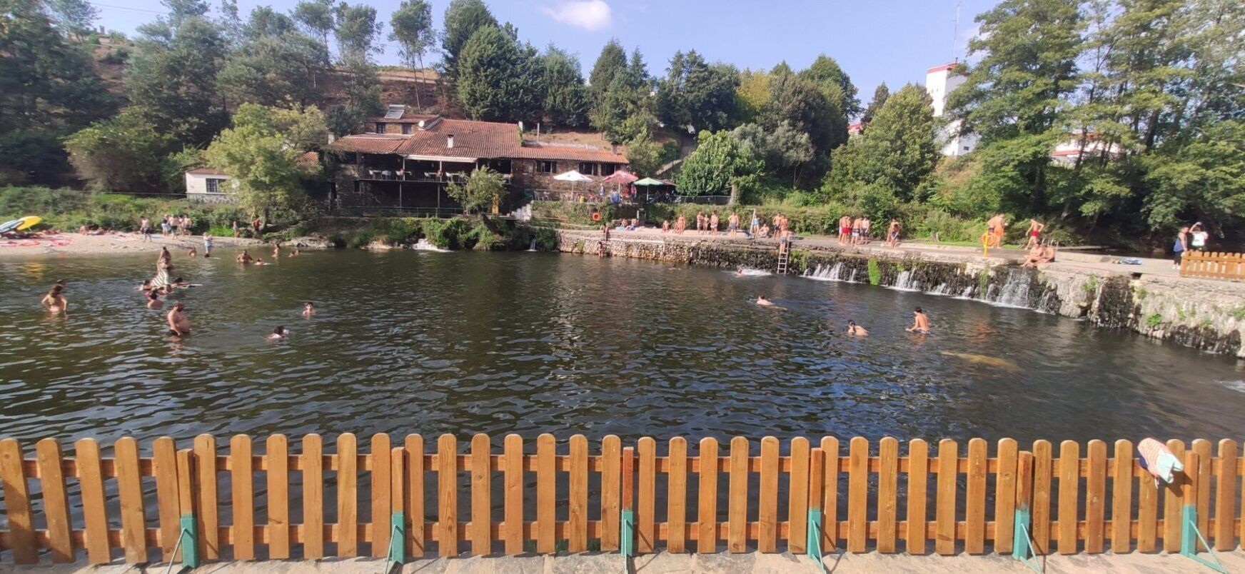 A swimming area with a wooden fence, a building with a tiled roof, and trees in the background.