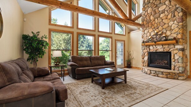 A spacious living room with a stone fireplace, large windows, and a high ceiling with exposed beams. - 3BR Townhome | Beach Access & Pontoon Rentals (Nisswa)