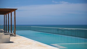 Una piscina con barrera de vidrio, terraza de madera y sillones de descanso.