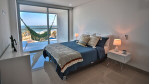 Un dormitorio con cama, mesita de noche, lámpara y vistas al océano a través de amplias ventanas.