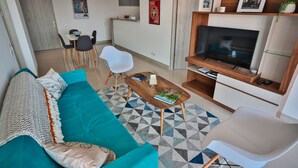 Una sala moderna con un sofá verde azulado, una mesa de centro y un televisor. Se observa un comedor con sillas y una mesa al fondo.