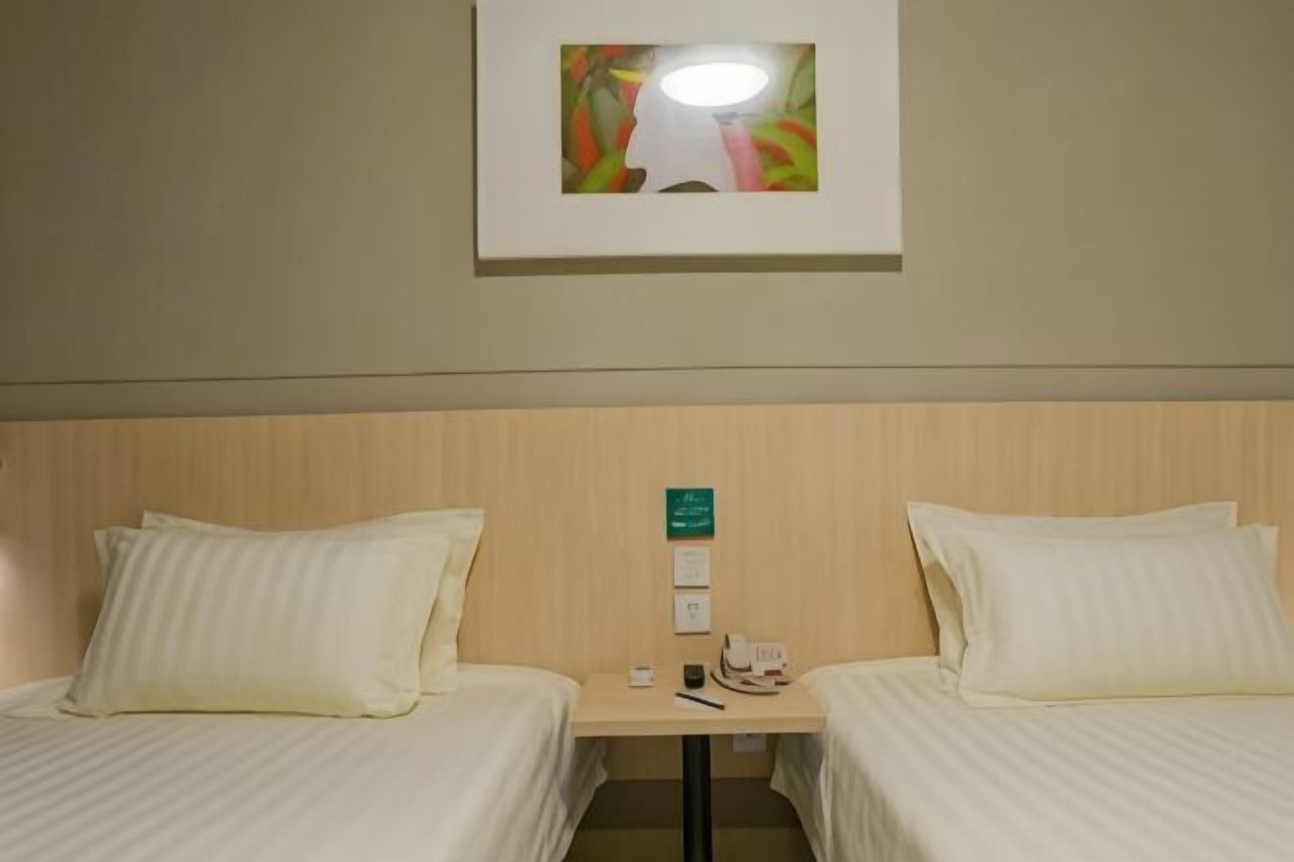 Habitación de hotel con dos camas, una mesita de noche, una lámpara, un cuadro enmarcado y un termostato fijado a la pared.