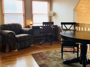 Um quarto aconchegante com um sofá xadrez, uma mesa de jantar de madeira e uma cadeira. Há um abajur em uma mesinha de cabeceira e uma janela com persianas.