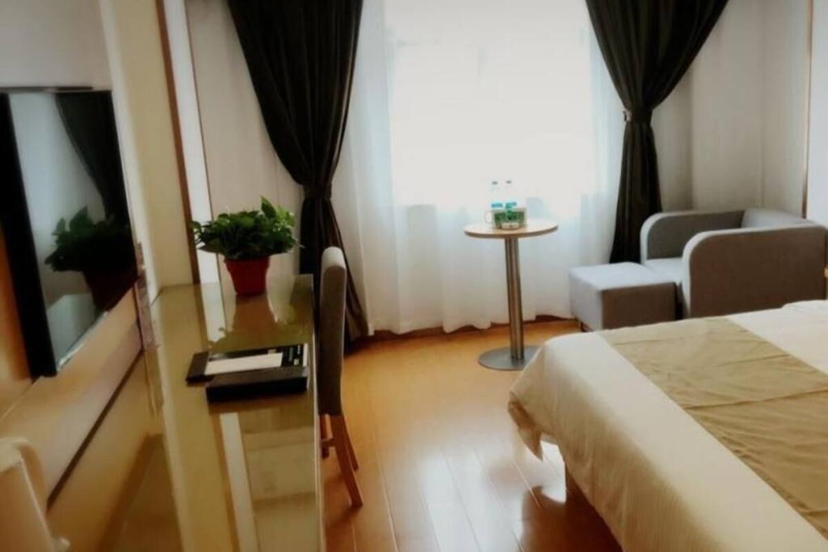 A hotel room with a bed, a desk with a chair, a small table with a plant, and a mirror.