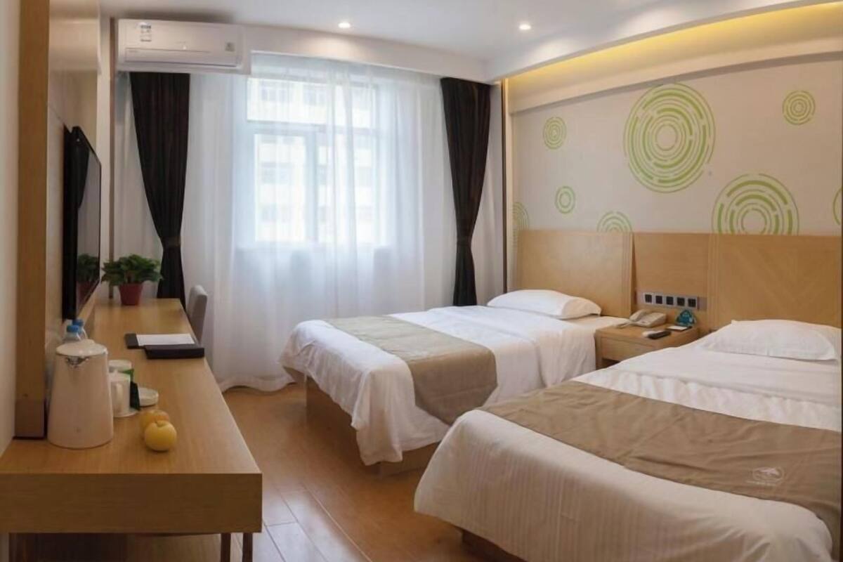 A hotel room with two beds, a desk with a chair, an air conditioning unit, and a window with curtains.