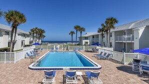 A swimming pool with blue chairs and umbrellas, surrounded by palm trees and a view of the ocean.
