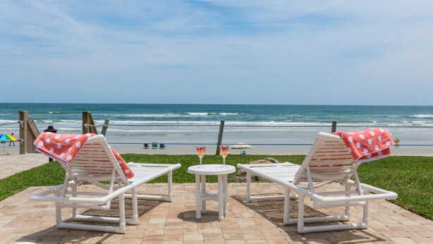 Two white lounge chairs with red polka dot towels, a small round table with two glasses of a drink, and a view of the ocean.