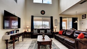 A living room with a large window, a flat-screen TV, a wooden coffee table, a brown leather sofa, and a dark-colored rug. - Dreams come true (Kissimmee)