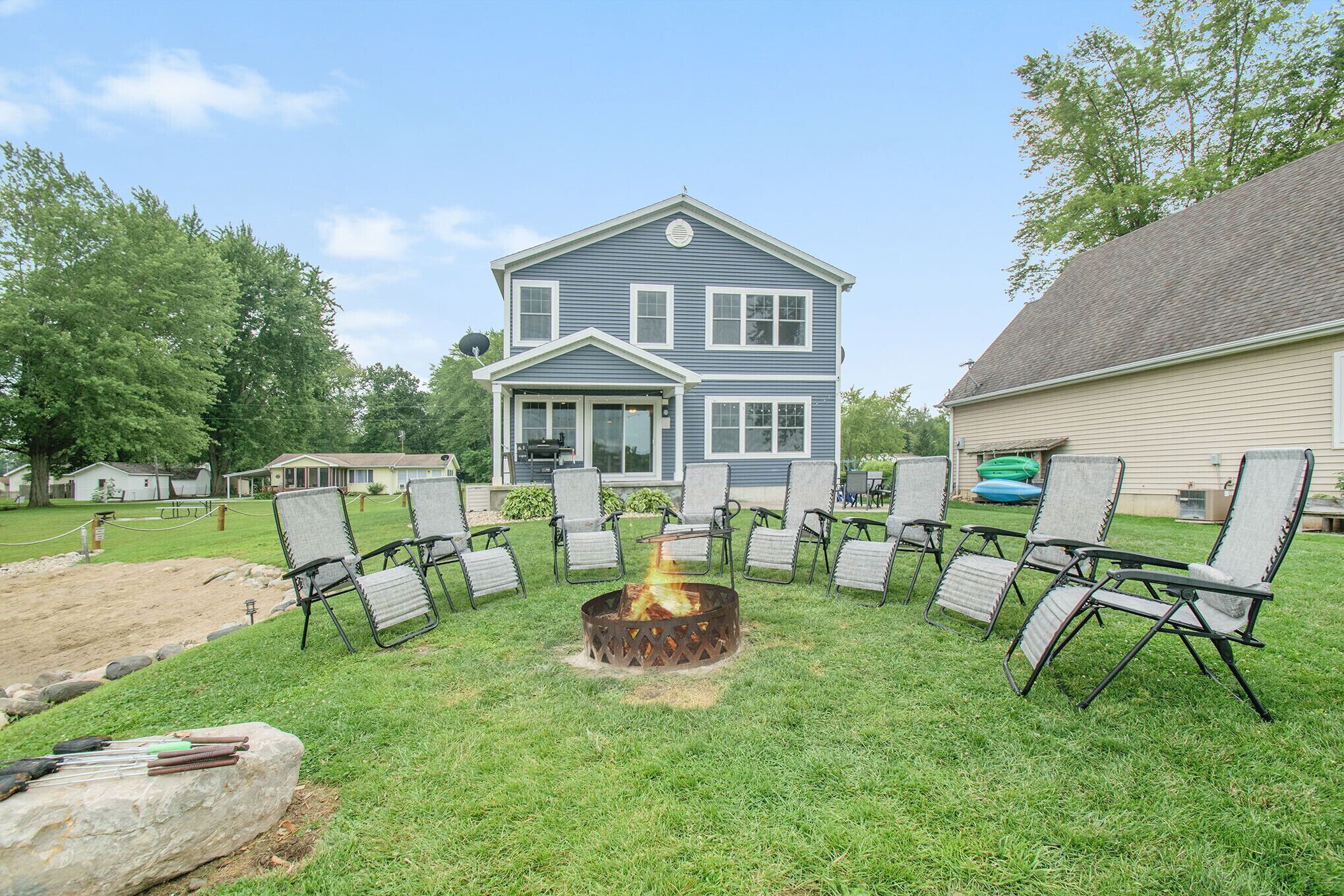 A backyard with a fire pit surrounded by lawn chairs, a house in the background, and a grassy area.