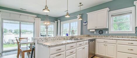 A spacious kitchen with white cabinets, a granite countertop, and a large island. There are multiple windows providing natural light and a view of the outdoors.