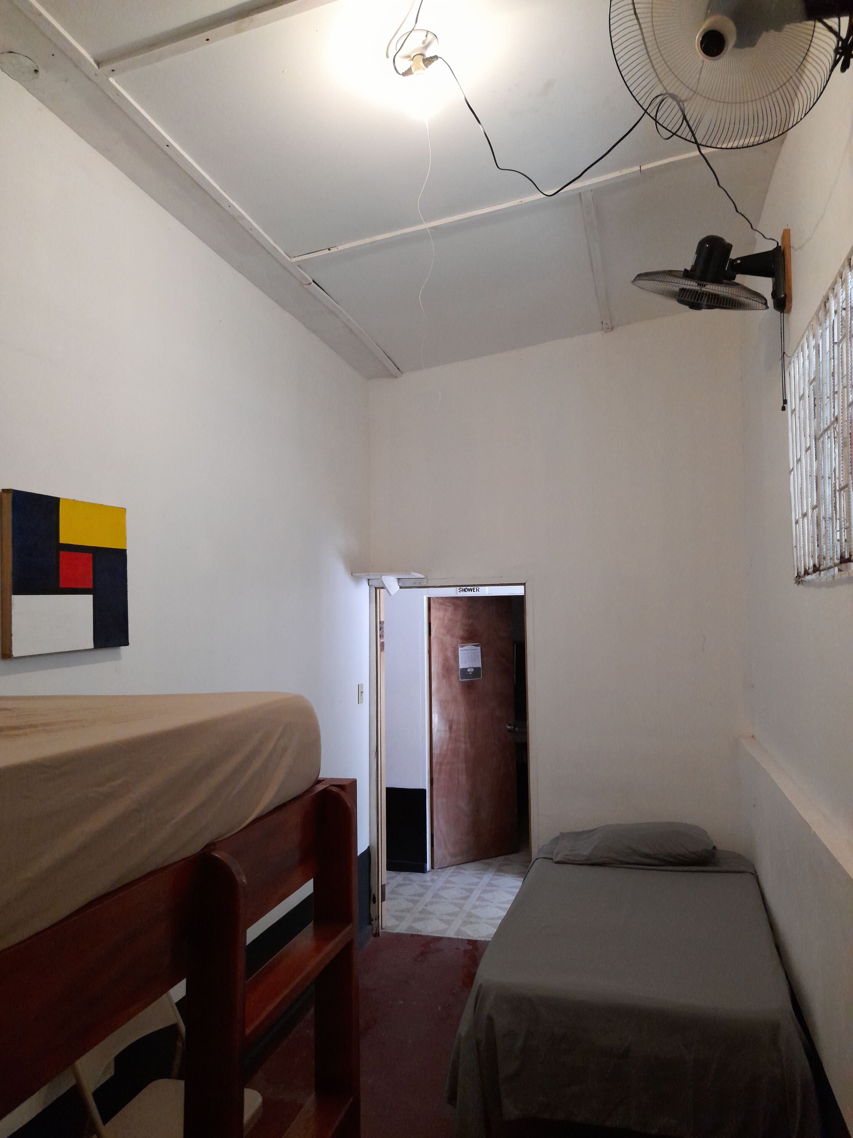 Una habitación pequeña y poco amueblada con una cama individual, una silla y un ventilador. Se observa una puerta de ducha al fondo.