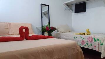 Habitación de hotel con dos camas, una con una toalla con forma de corazón y la otra con una toalla amarilla. Hay un espejo en la pared y un pequeño estante con un ventilador y un televisor.