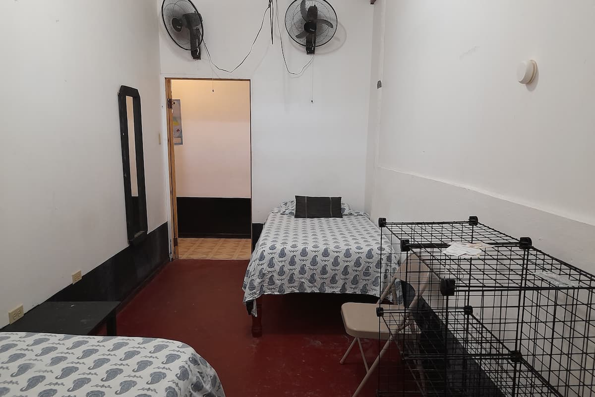 A small, compact room with two beds, a desk, and a chair. There are two wall-mounted fans and a door leading to another room.