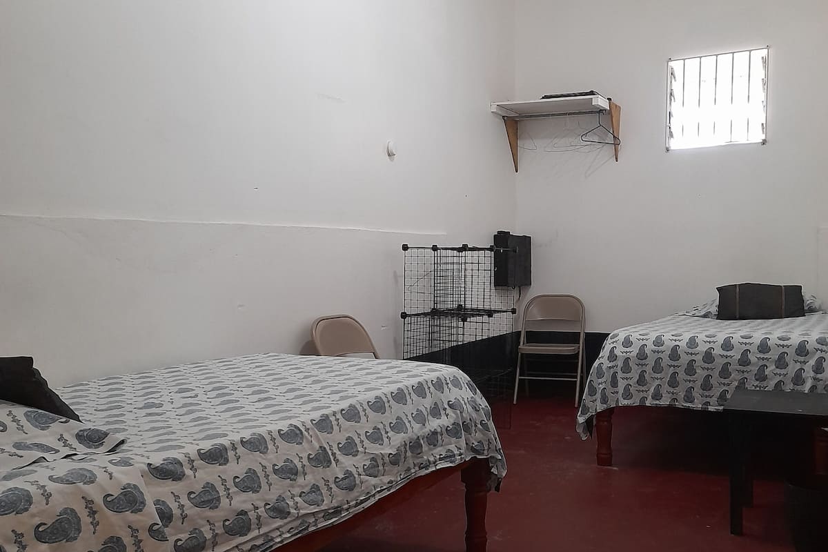 A room with two beds, a desk, a chair, a small window, and a wall-mounted shelf.