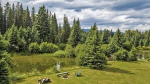 A serene outdoor setting with a grassy area, a bench, and a small fire pit surrounded by dense forest and mountains in the background. - 1920 Montana Quiet Log Cabin! Creek, fire pit, 10 ac. of wilderness/ Swan Valley (Bigfork)