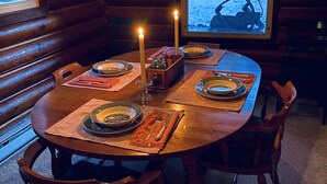 A cozy cabin interior with a wooden dining table set for four, candles, and a window with a view of a snowy landscape. - 1920 Montana Quiet Log Cabin! Creek, fire pit, 10 ac. of wilderness/ Swan Valley (Bigfork)