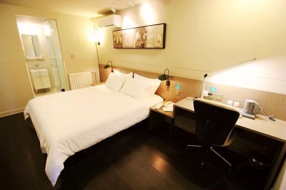 A hotel room with a bed, bedside table, chair, desk, lamp, and a painting on the wall.