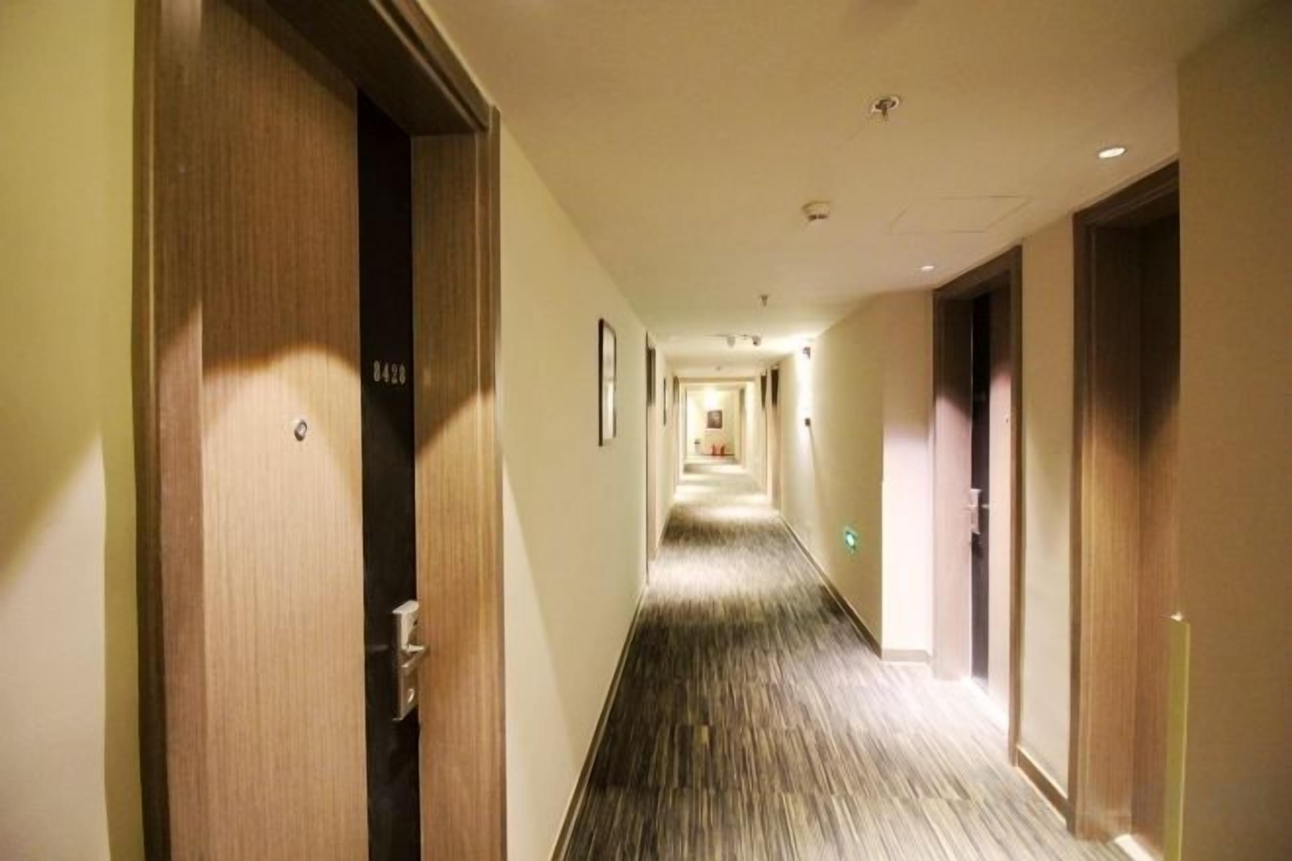 Corredor de hotel com portas de madeira, piso revestido de carpete e um longo hall que leva a uma área comum.