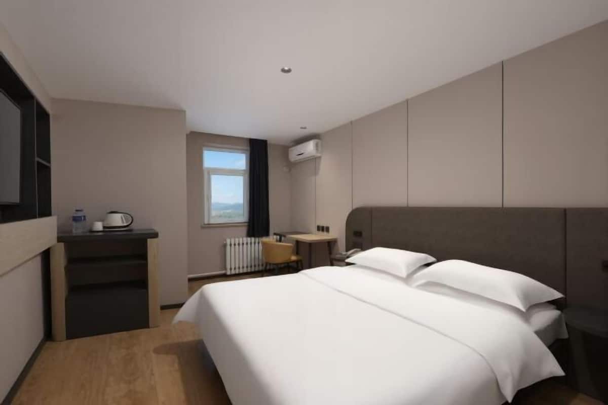 A hotel room with a bed, a desk, a chair, a radiator, and a window.